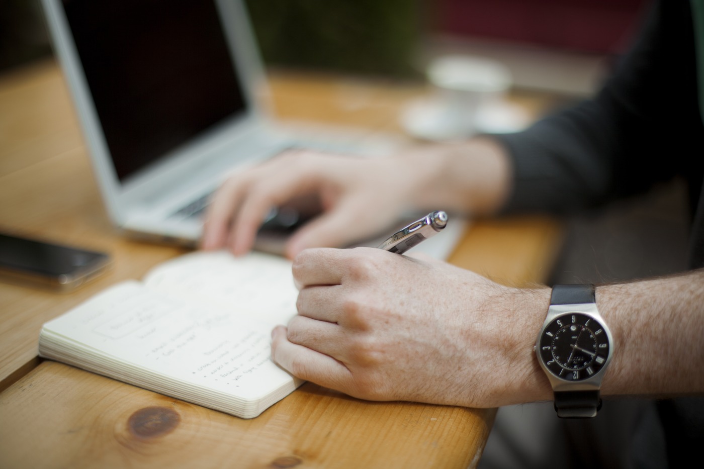 Hands writing notes in a notebook beside an open laptop